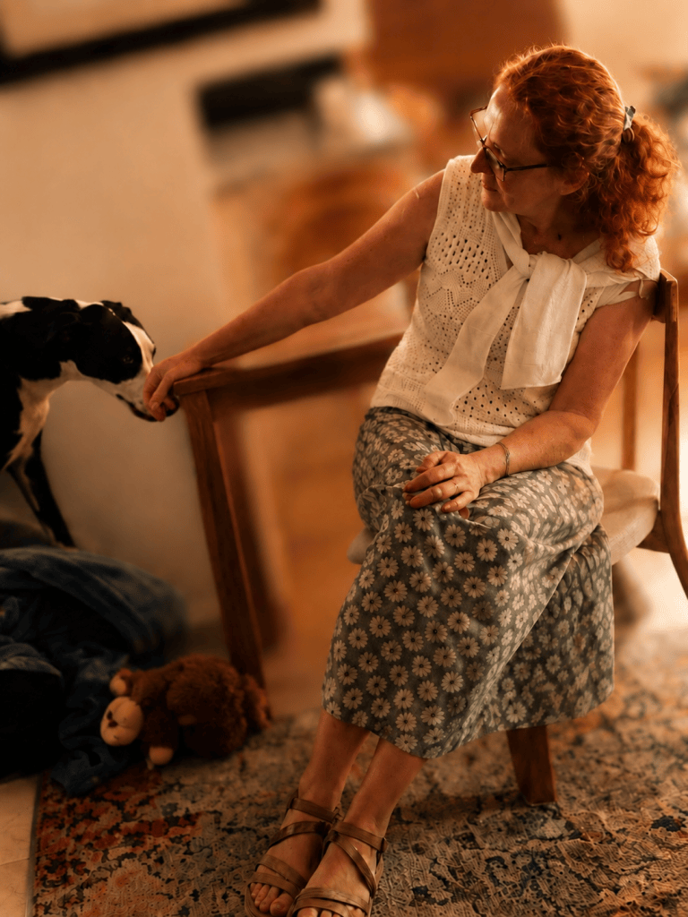Claire Baley seated at home, gently reaching out to Bea the dog in warm evening light.
