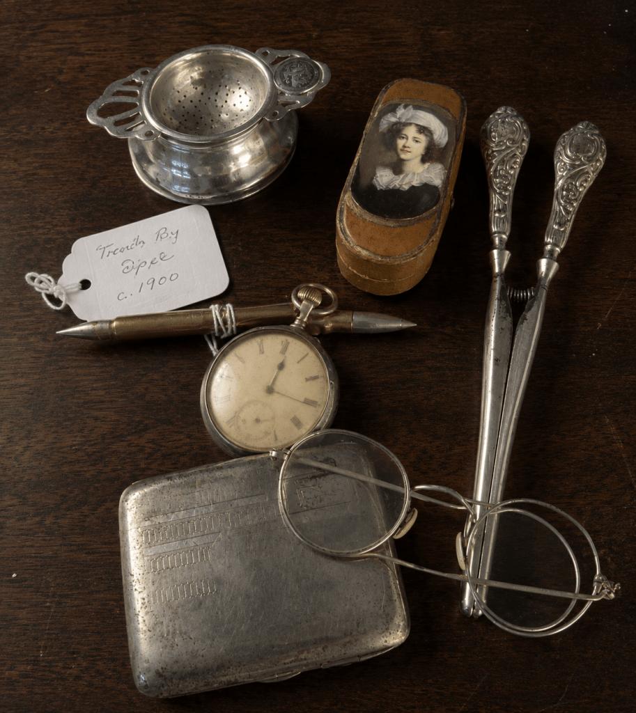 Collection of small antique objects including a pocket watch, spectacles, silverware and keepsakes arranged on a dark wooden table.